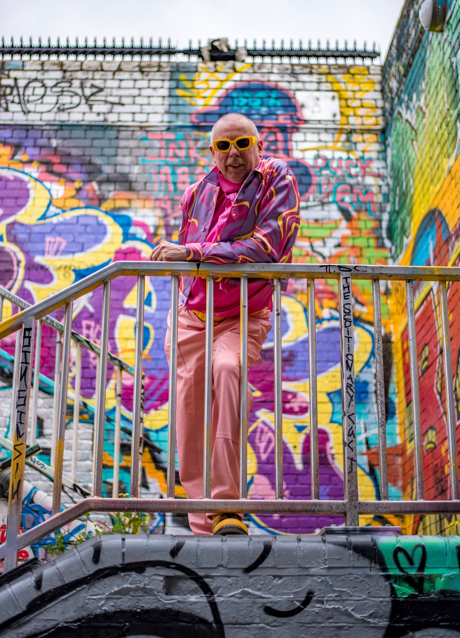 Gentleman wearing pink trousers, pink shirt and pshychadelic shirt standing against colourful wall
