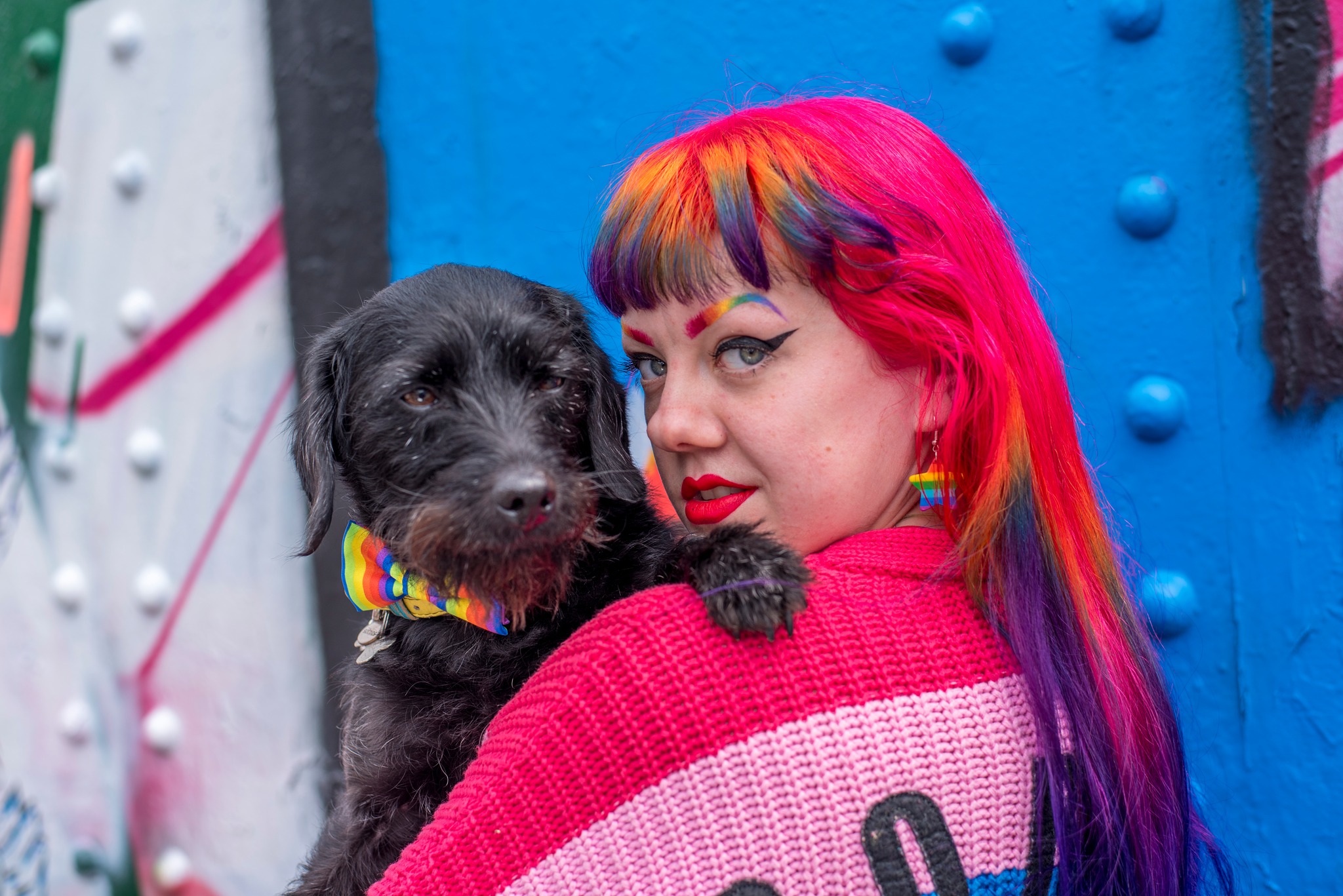 Girl with rainbow hair and cardigan holding a black dog wearing a rainbow bowtie