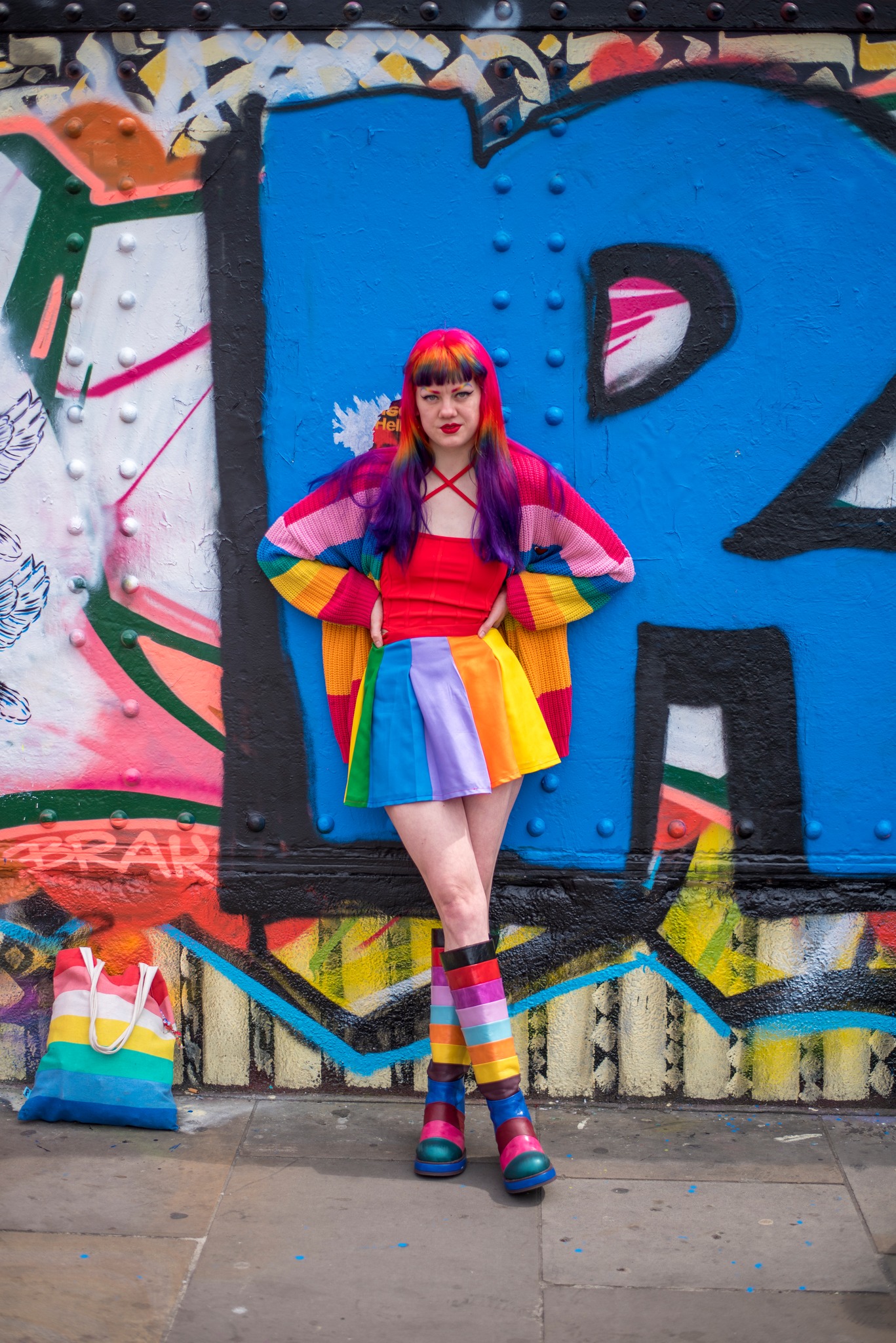 Grace has rainbow hair and wears a rainbow skirt and boots standing against a colourful wall
