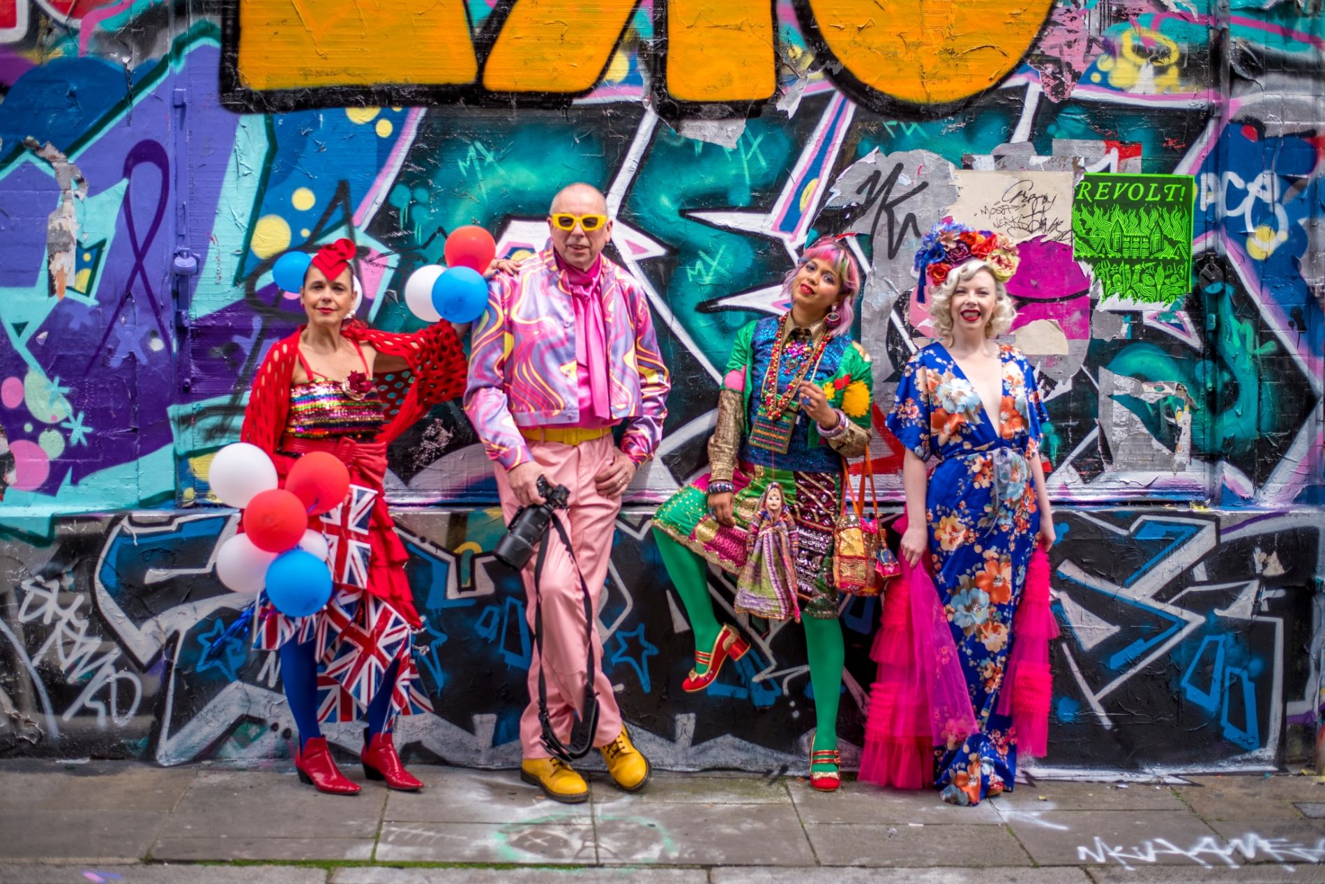 4 colourfully dressed people standing against a wall covered in spray can street art