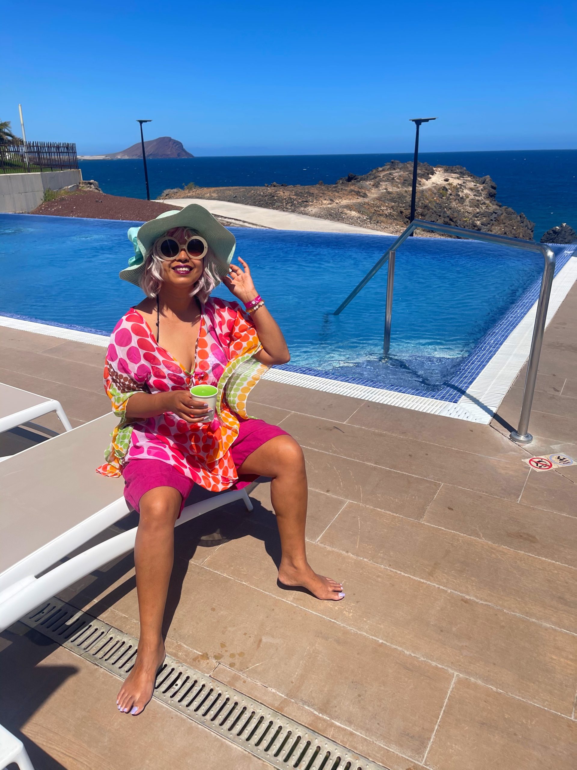 girl wearing sunglasses and sun hat holding a green slush drink sat on lounger with pool behind