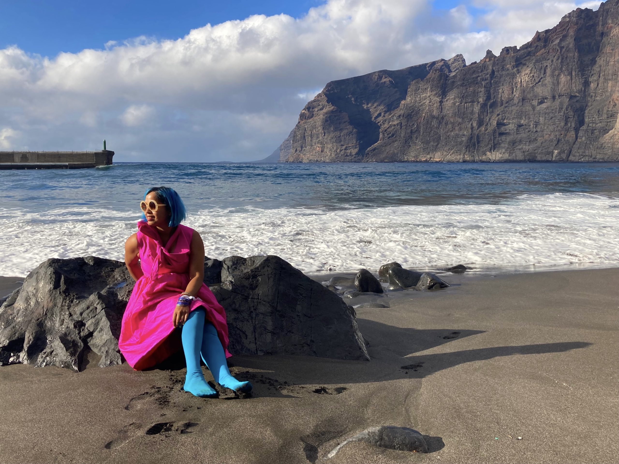 girl in pink dress sitting against a rock on black sand beach with sea in background