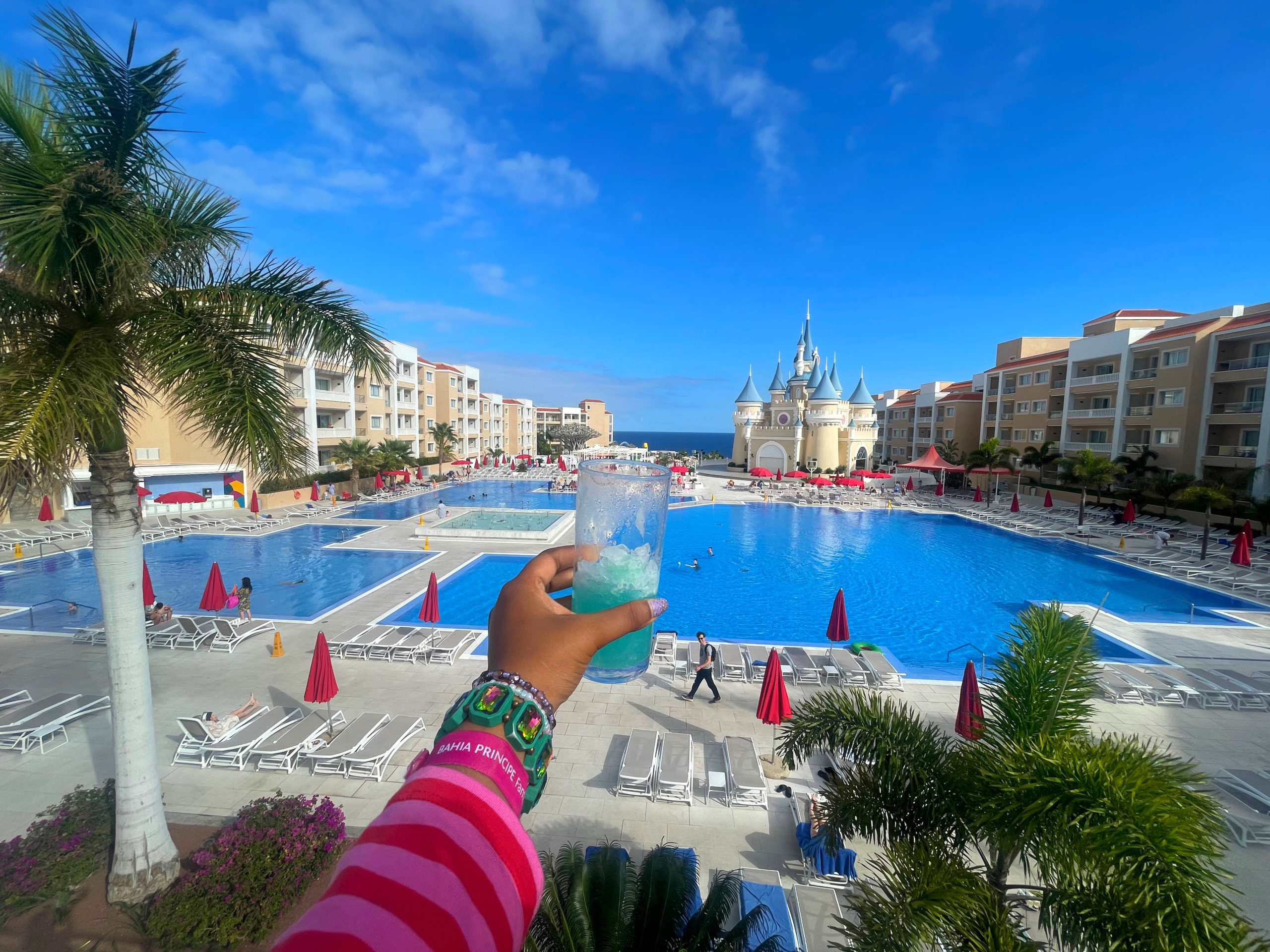 hand holding a cocktail over the view of the bahia principe fanastia tenerife colourful hotel - you can see the pool and castle in view