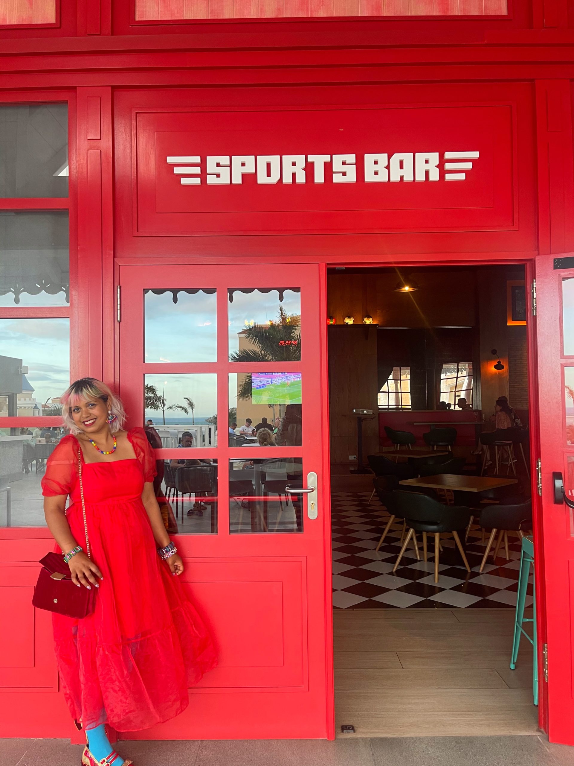 Momtaz wears red dress and stands out side a red bar with sports bar written on it