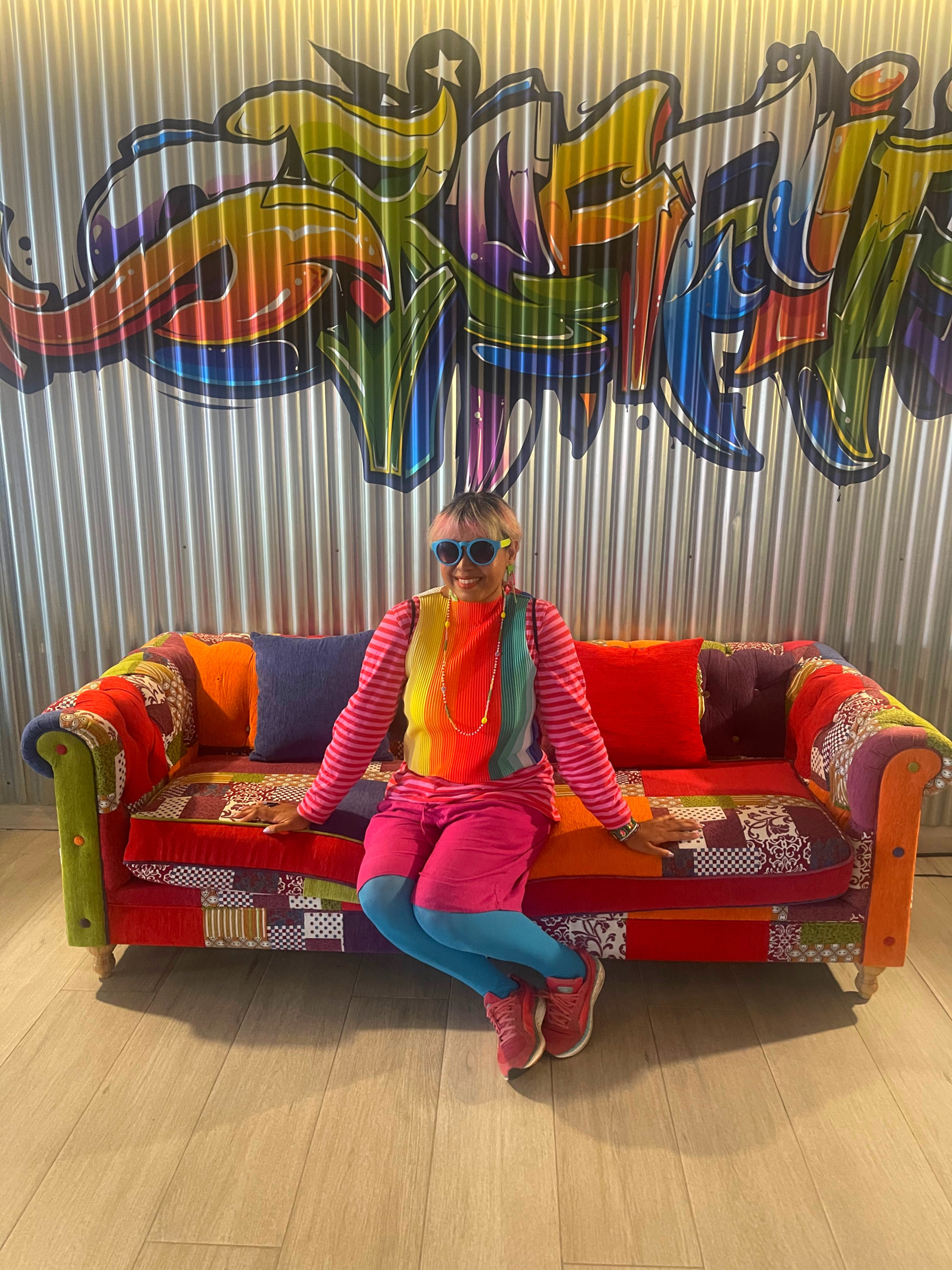 colourful hotel tenerife, colourfully dressed girl with sunglasses sat in centre of a patchwork sofa with metal wall behind with spray can art on