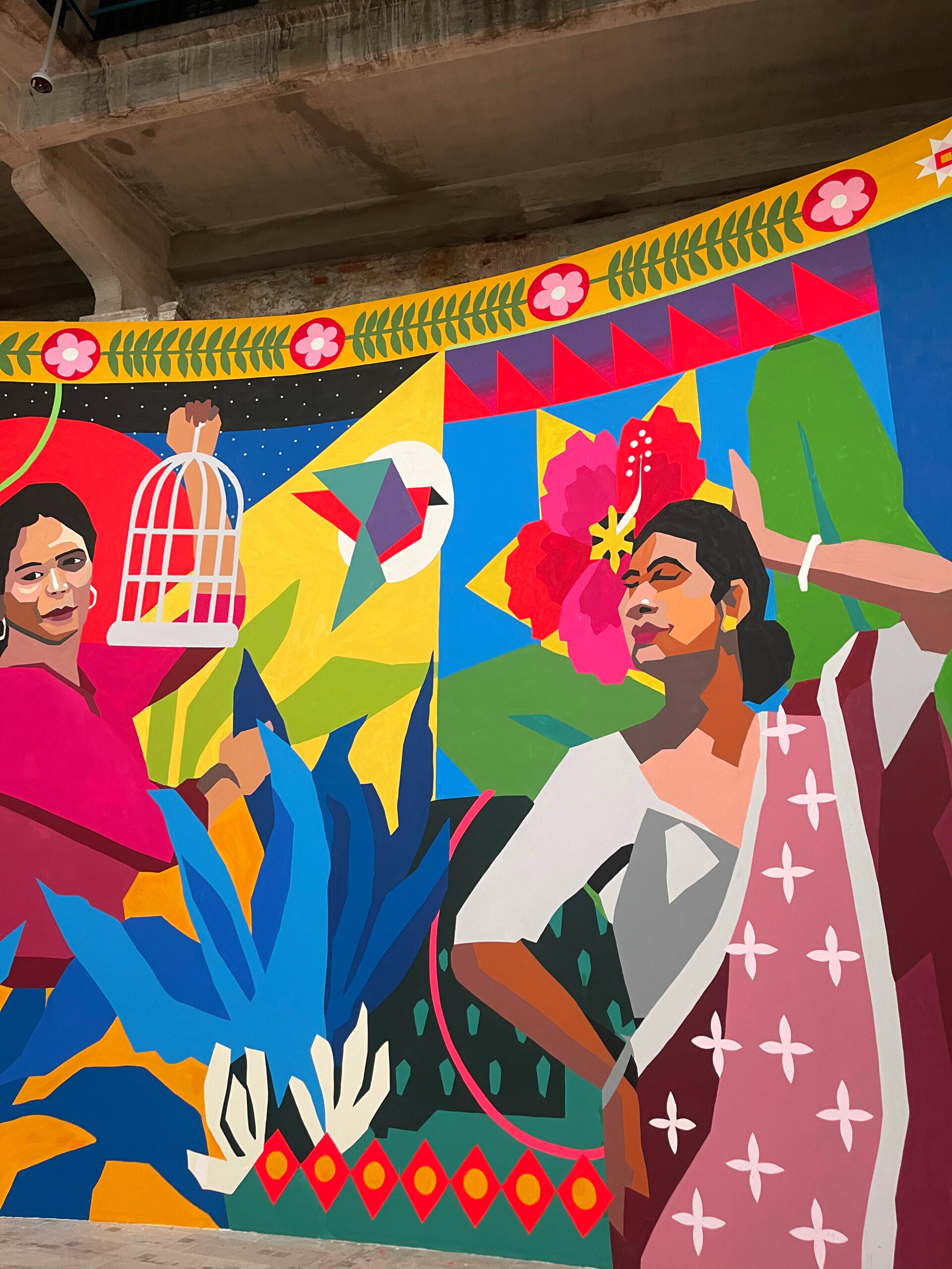 a large painted mural celebrating the trans community in india. it's painted in blod block colours and looks like an illustration of a transperson dancing in a sari and one holding a  birdcage