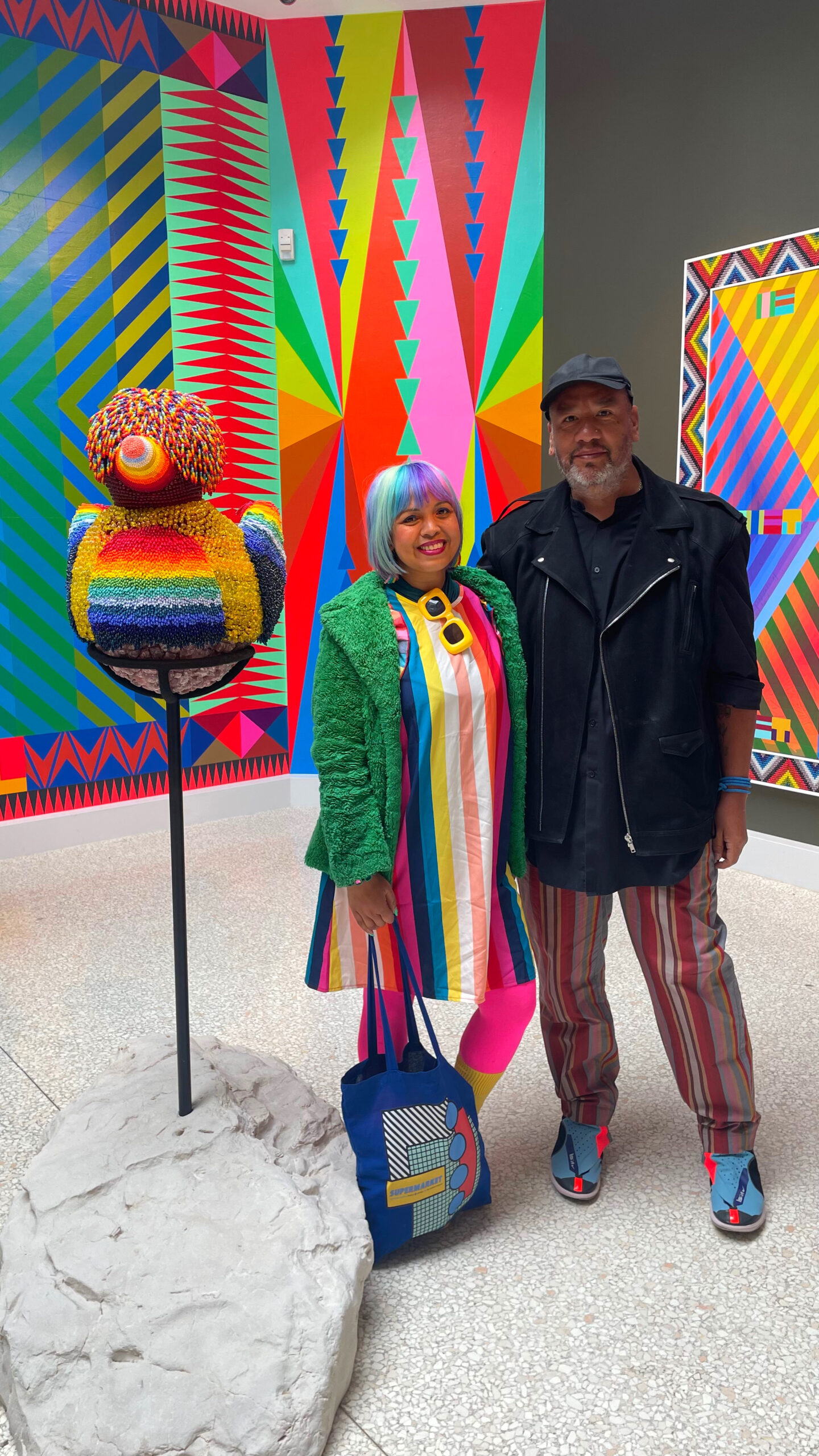 writer standing next to the artist, she is wearing a stripey dress and he has stripey trousers they stand next to his beaded duck