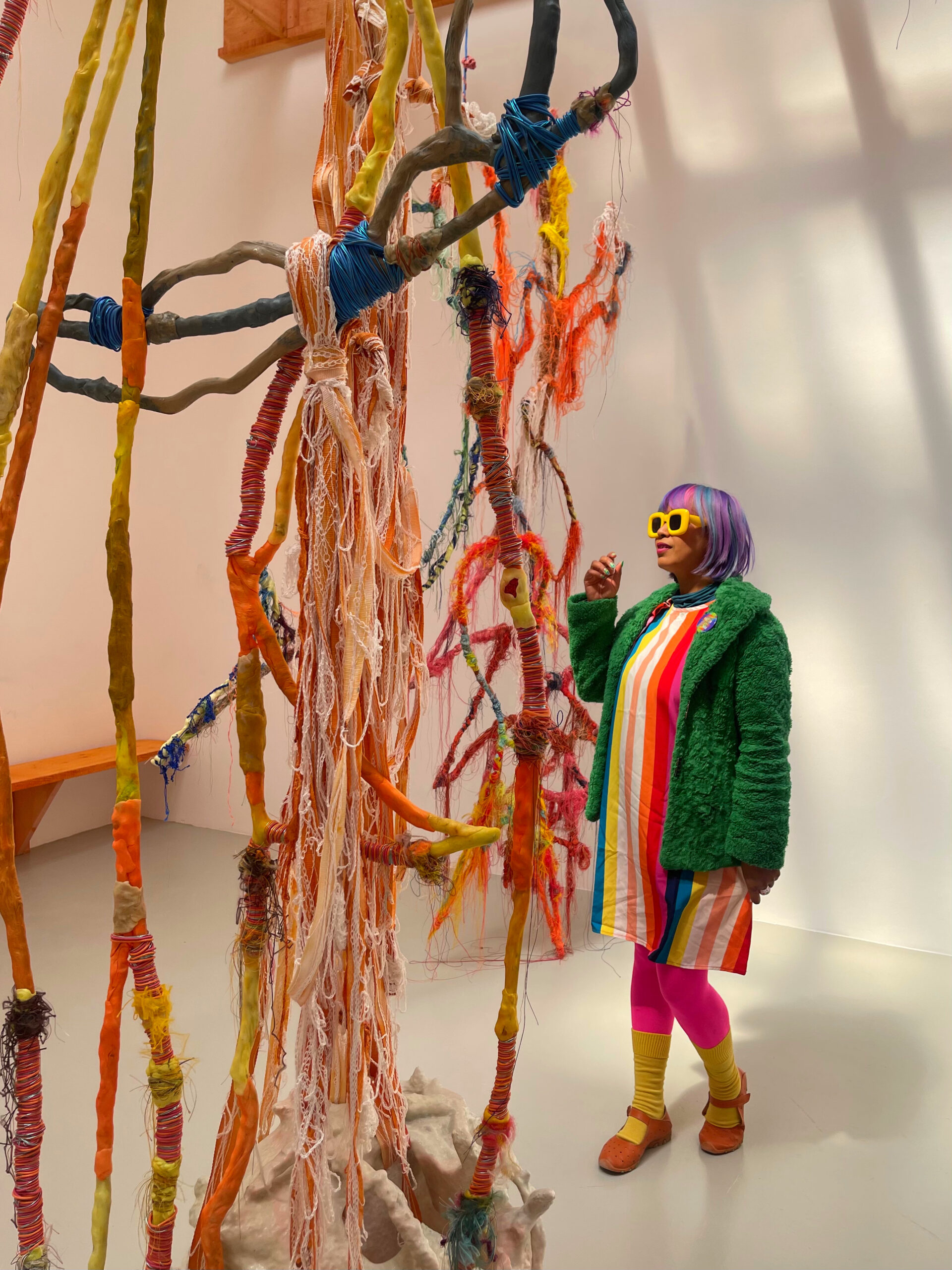 a series of what looks like branches of a tree covered in colourful wax and yarns in a white room, a girl in a green coat and yellow sunglasses looks up at it