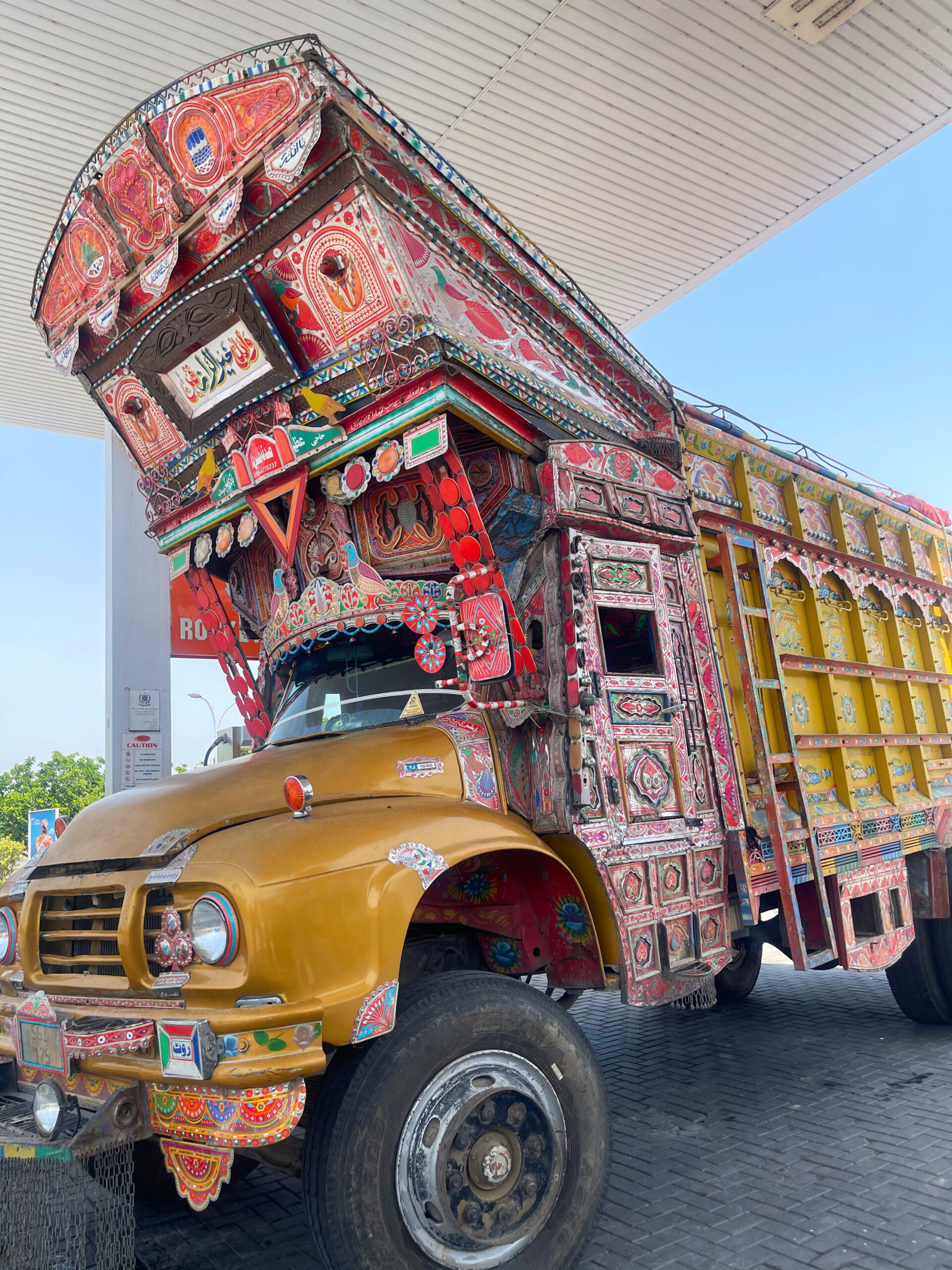 a large yellow based old style of pakistani truck art with wooden hood parked at a service station
